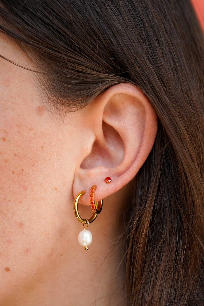 Close-up of a person's ear wearing a gold huggie hoop earring with a dangling white pearl and a small orange beaded accent, alongside a small red stud earring.