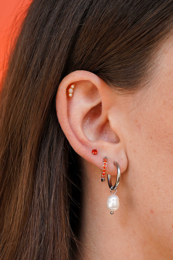 A close-up of a person's ear showcasing multiple earrings, including a silver huggie hoop with a dangling white pearl, a small orange gem stud, a silver and orange gem hoop, and a beaded cartilage flat back stud.