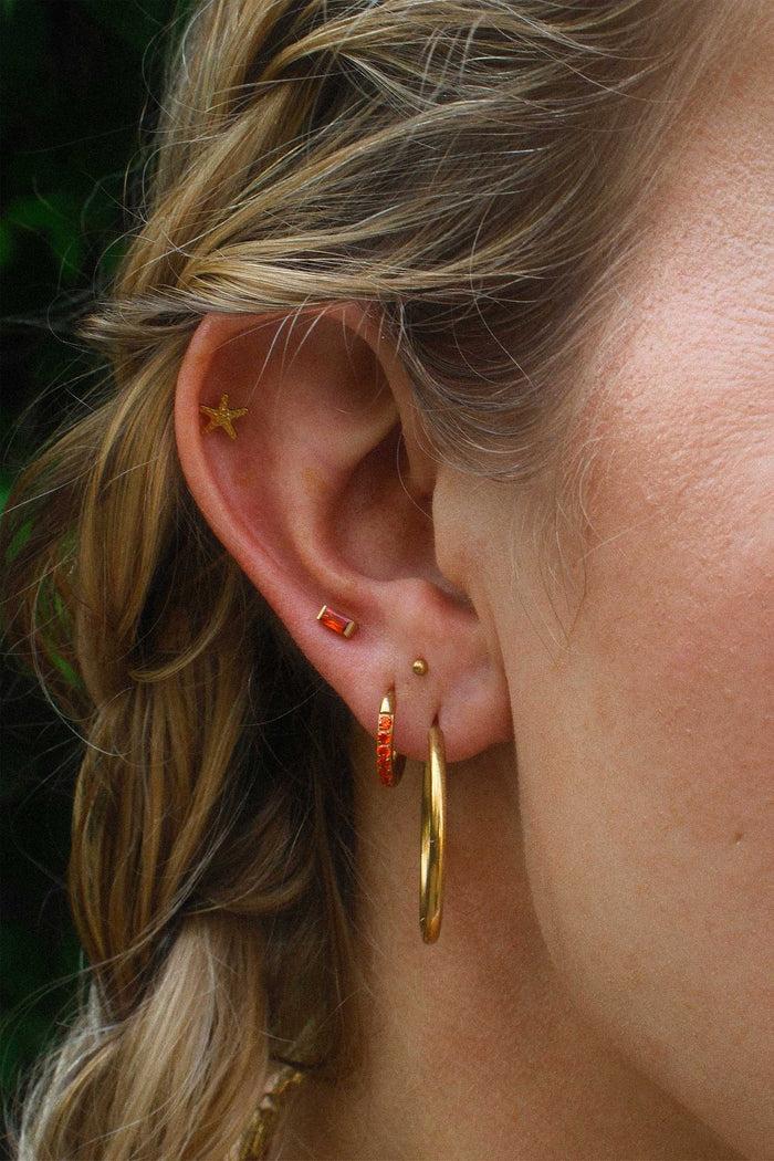 Close-up of an ear with gold hoop earrings and orange studs against a blurred background.