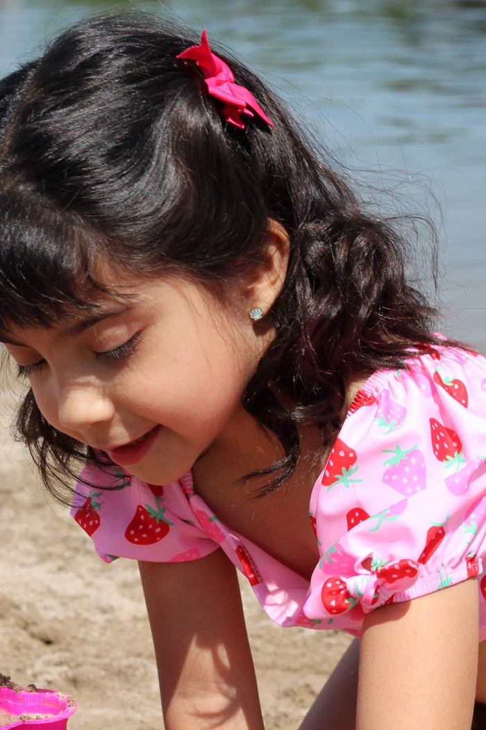 Young girl with a blue shell-shaped earring in a pink swimsuit playing by a body of water.