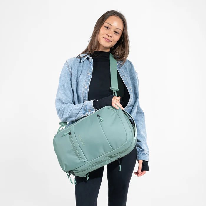 A woman wearing a Sherpani Camden Surf Blue Ultimate Backpack with crossbody strap against white background. #color_surf-blue