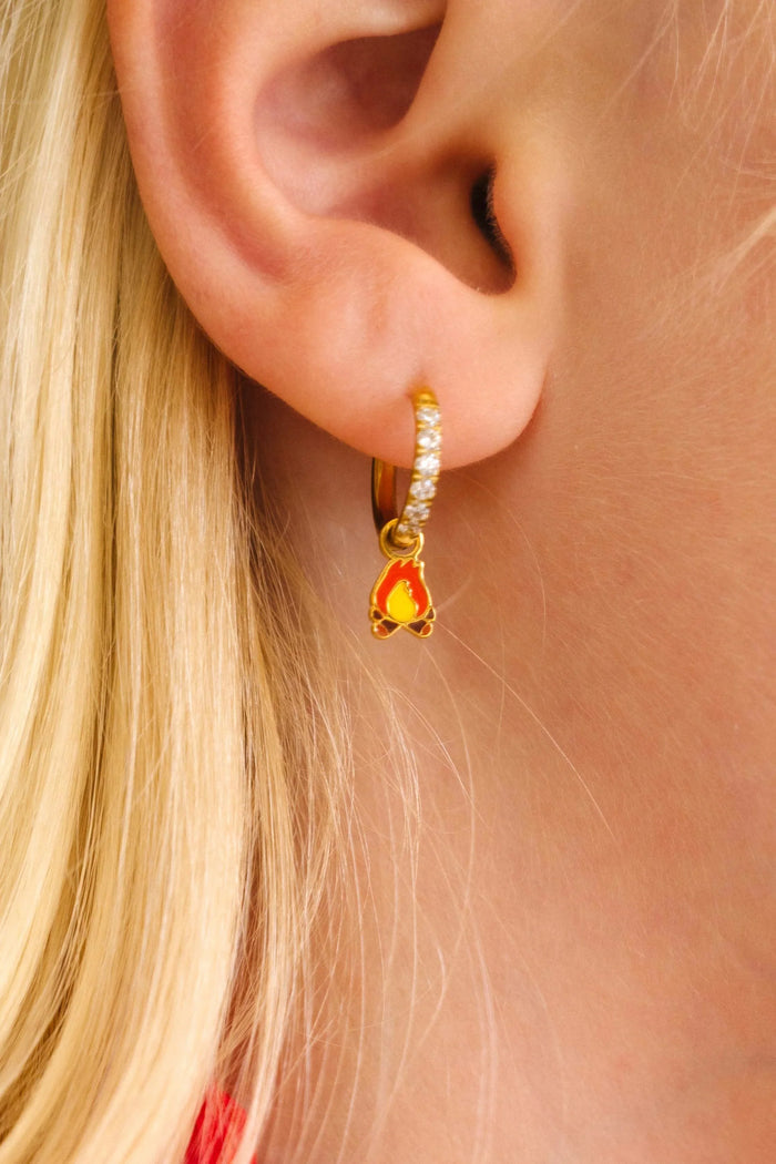 Close-up of a person wearing gold hoop earrings with enamel campfire charm, a red bandana, and a white shirt.