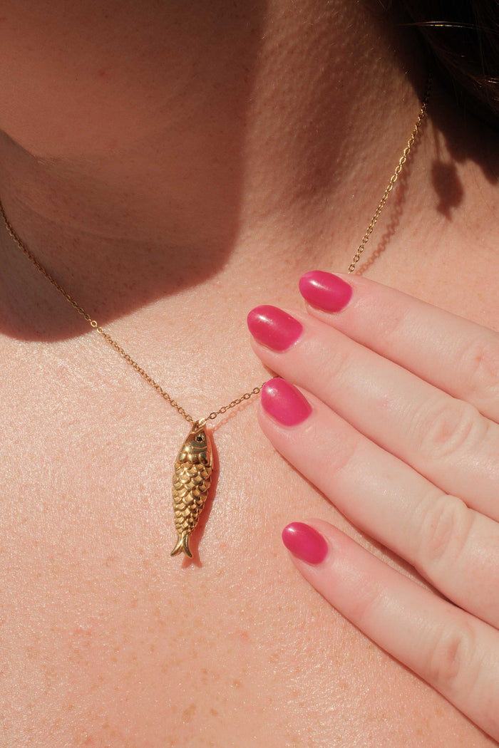 Gold necklace with a fish pendant worn by a person with pink nail polish.