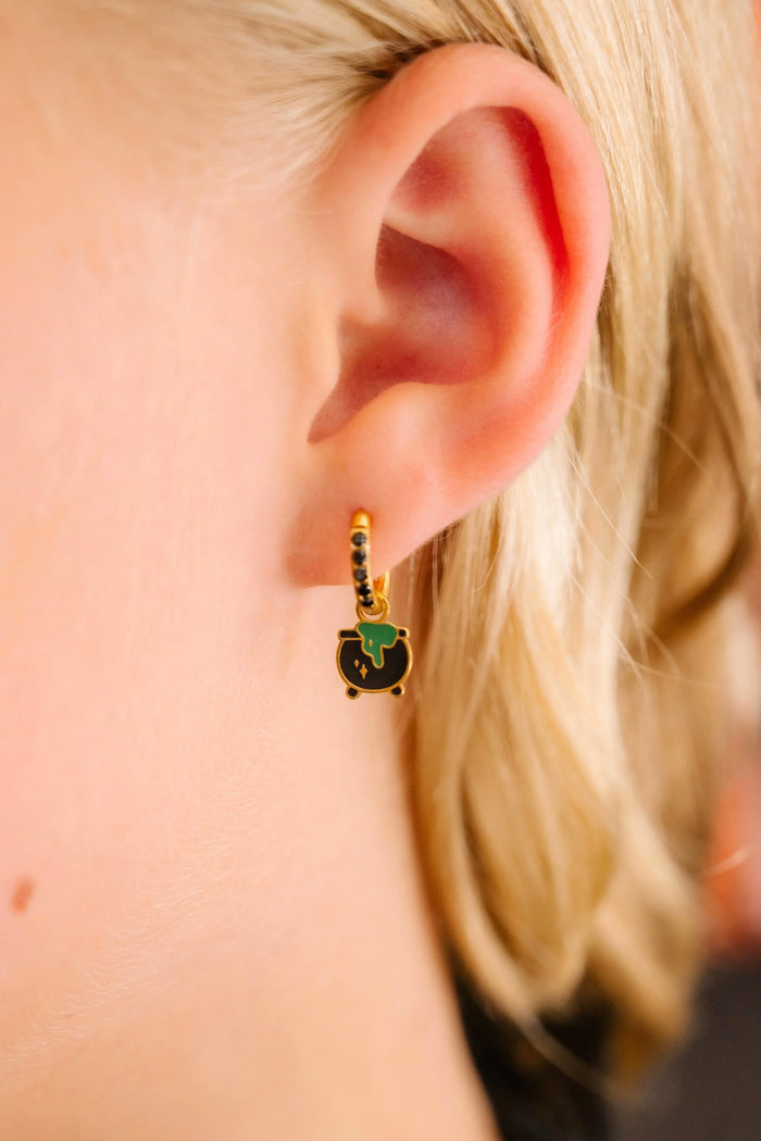 Close-up of an ear wearing a gold earring with green and black enamel charm shaped like a cauldron.