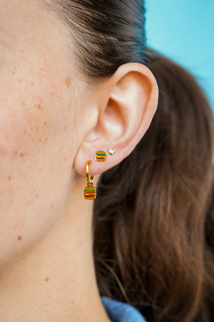 Close-up of an ear wearing a gold cheeseburger-shaped flat back stud earring and a gold hoop earring with a blue background