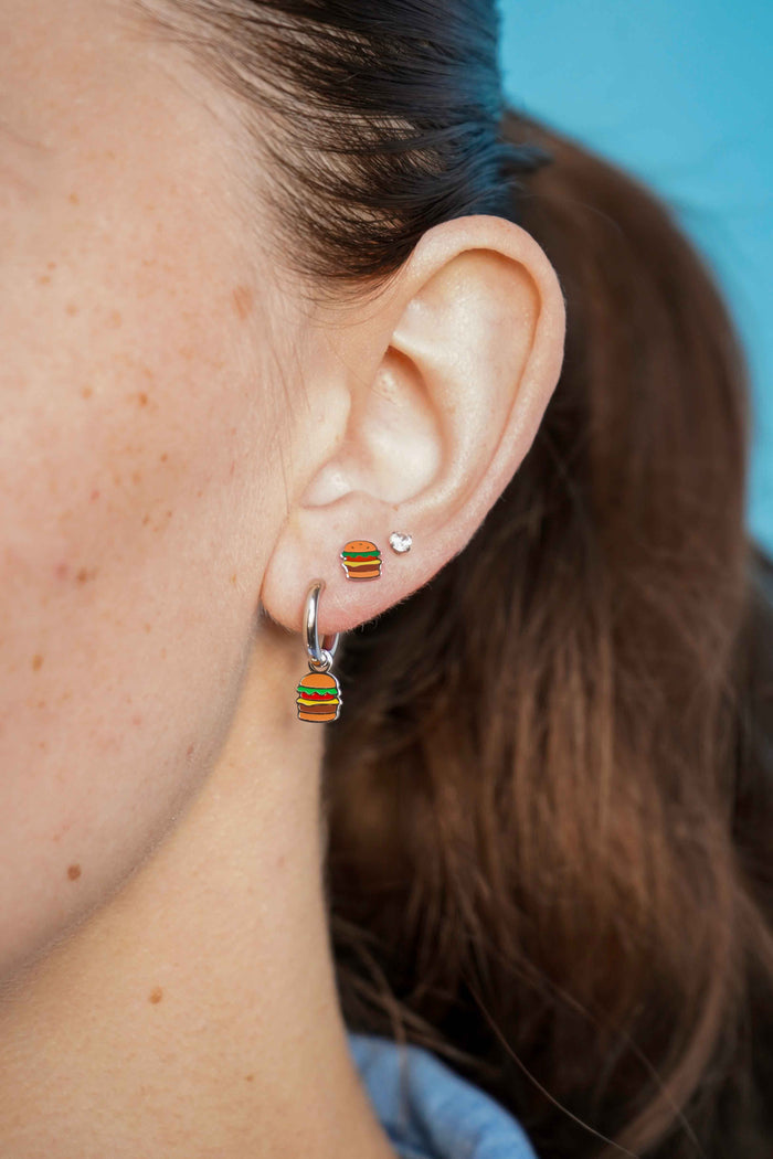 Close-up of an ear wearing a silver cheeseburger-shaped flat back stud earring and a silver hoop earring with a blue background