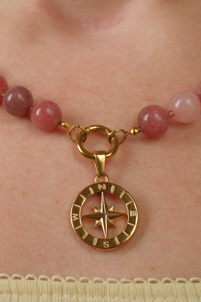 A close-up of a beaded necklace worn on a person's neck, featuring round pinkish-red beads and a gold compass pendant.