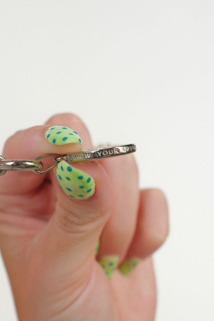 A close-up of a hand with green and yellow patterned nail art holding a silver chain and a side view of a silver compass pendant engraved with 'FOLLOW YOUR TRUTH'.