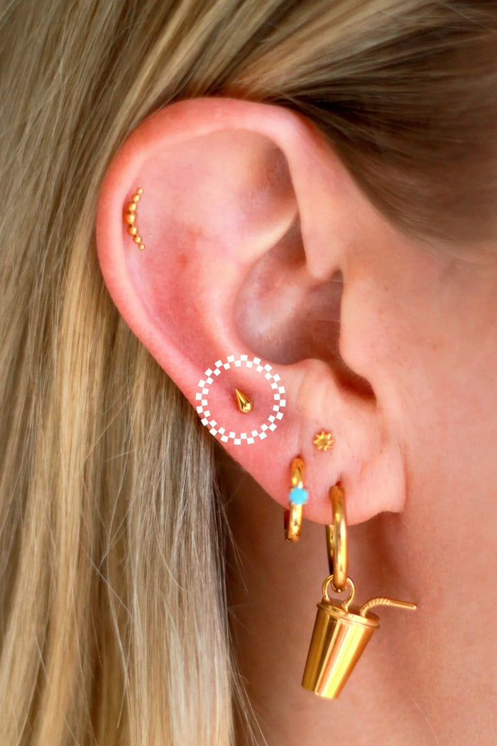 Close up of an ear with a lot of piercings, featuring gold flat backs studs and hoops. One flat back stud has been circled as the feature product in this listing. It is a gold, 3D teardrop shape.