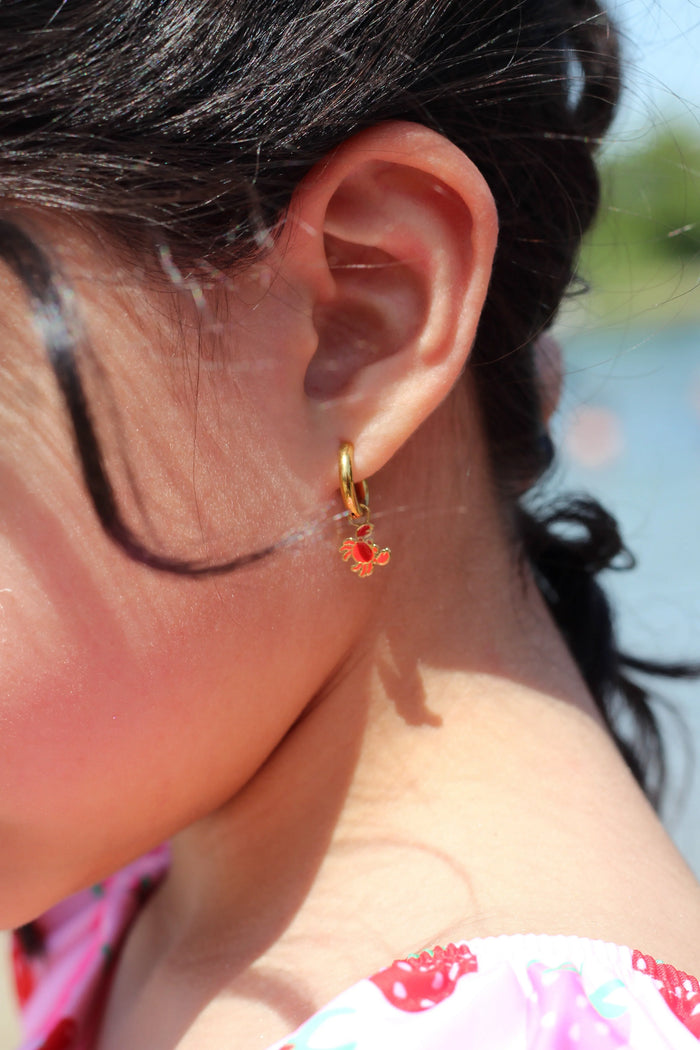 Close-up of a person's ear with a red crab charm on a gold hoop earring, outdoors.