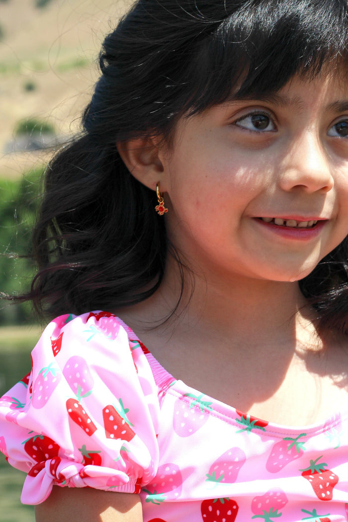 Young girl wearing a gold hoop with red crab charm on it and a pink shirt with strawberry pattern, outdoors