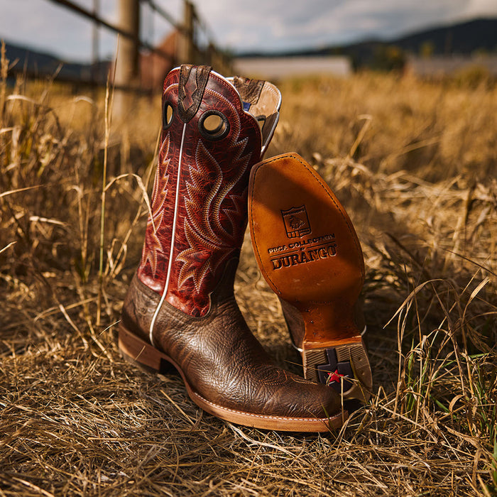 Brown cowboy boot with a red leather shaft on a grassy fields next to a fence.