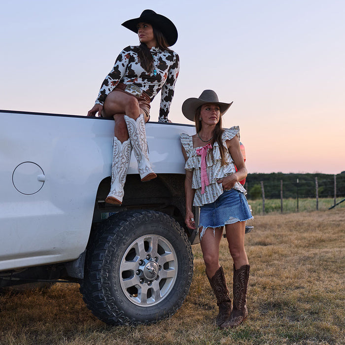 Two women in cowgirl hats and cowgirl boots standing next to a white truck in a field at sunset.