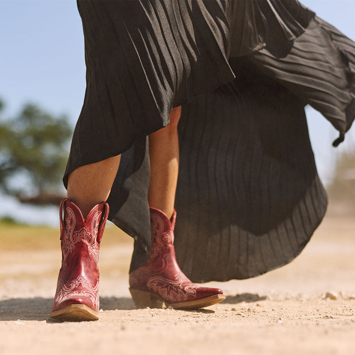 A person outdoors wearing a long flowing black skirt and red cowgirl boots, on a dirt path.