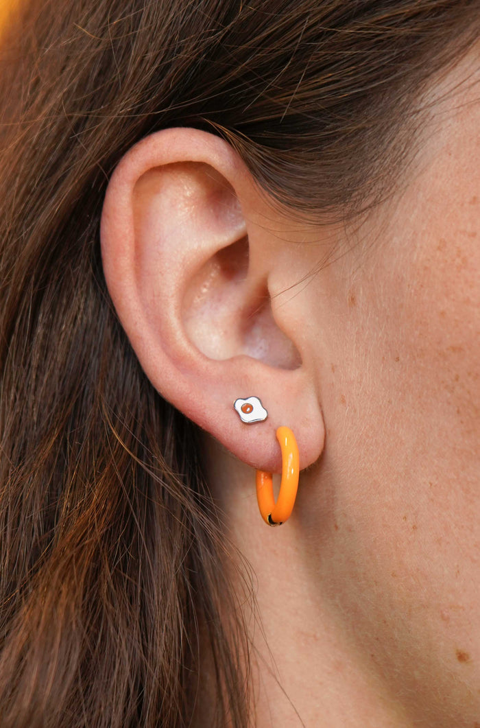 Close-up of an ear with a silver, white, and orange enamel flat back stud earring and orange hoop earring