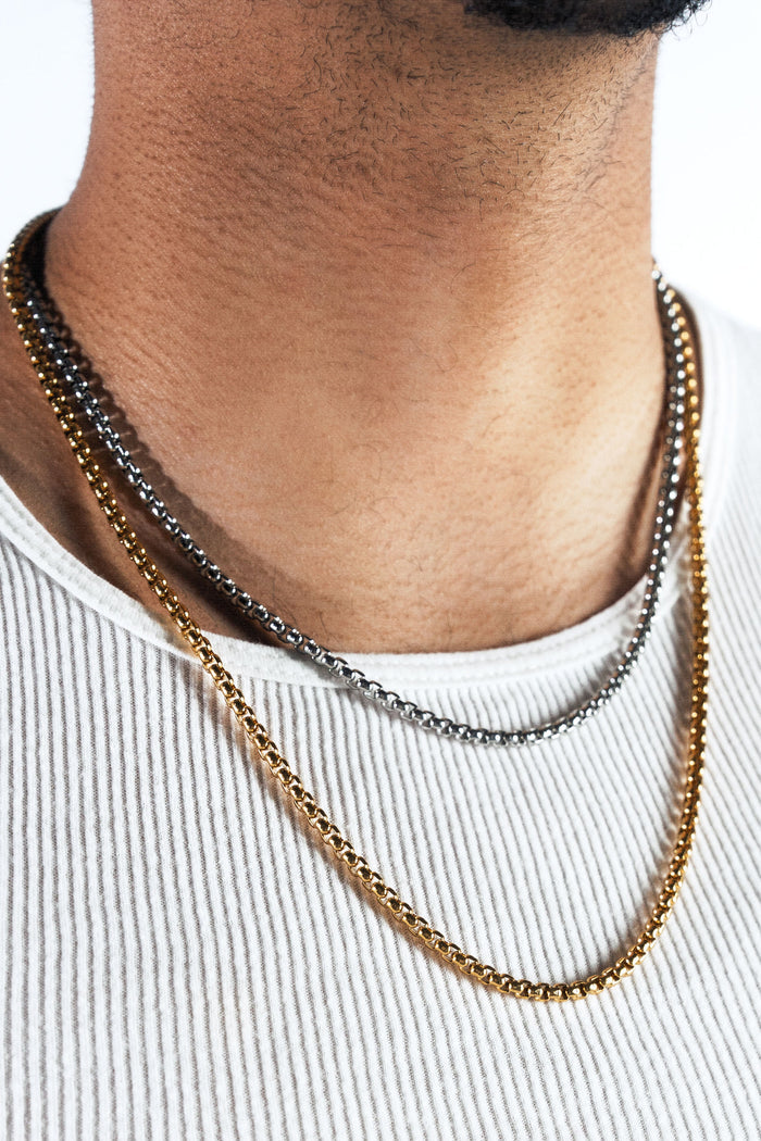 A close-up of a man's neck and chest wearing three layered chain necklaces: a gold curb chain and a silver curb chain, styled with a white ribbed tank top.