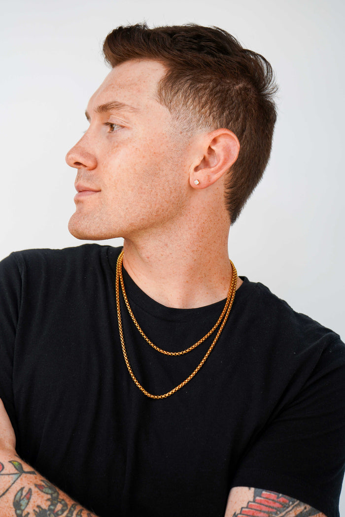 A man in a black t-shirt wearing two layered gold chain necklaces, shown in profile against a light gray background.