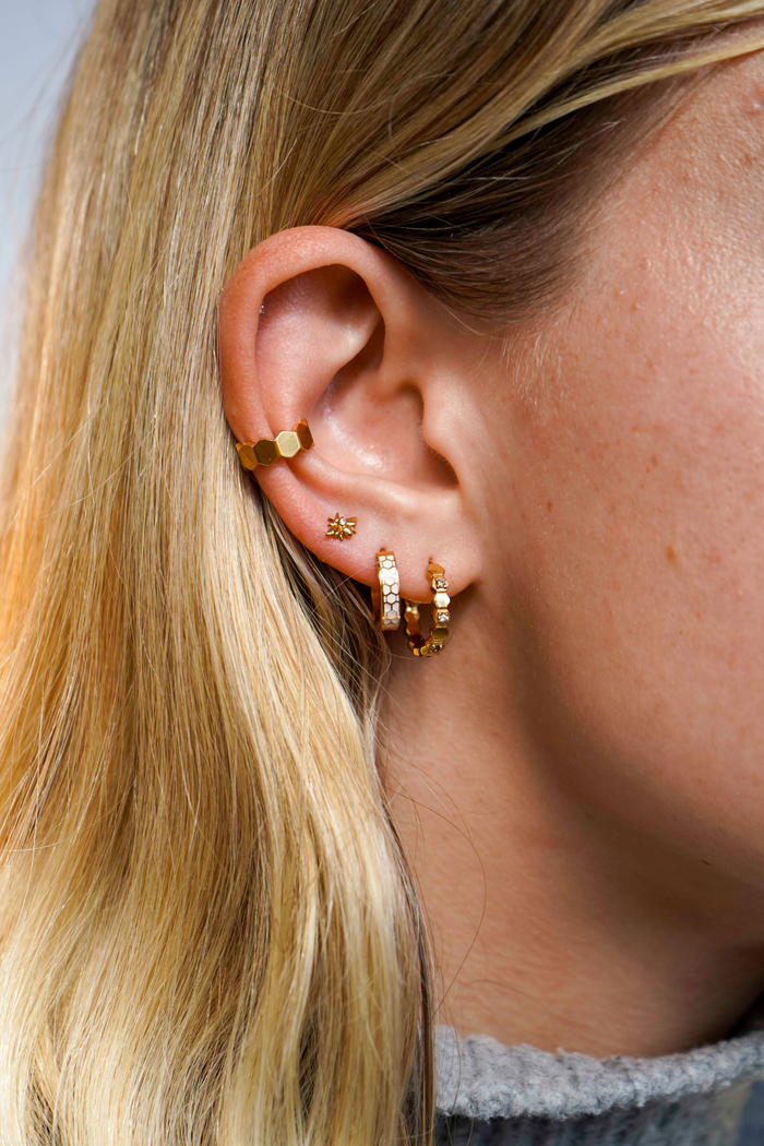 Close up of a model wearing a hoop earring made from stacked hexagon shapes with a honey colored zirconia stone set into every other hexagon shape. Hoop made from 18k gold-plated 316L Surgical Steel, , with a honey-colored zirconia stone, against a blurred background.