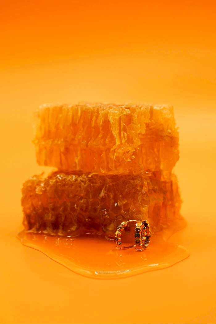 Close up of a model wearing a hoop earring made from stacked hexagon shapes with a honey colored zirconia stone set into every other hexagon shape. Hoop made from 18k gold-plated 316L Surgical Steel, ,with a honey-colored zirconia stone, sitting next to honeycomb and in a pool of honey, against a warm yellow-orange background