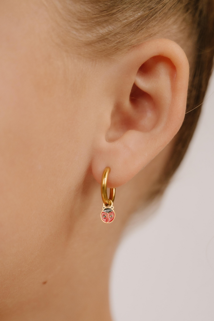 A close up of a ladybug shaped hoop charm with red and black enamel and gold-plated titanium metal hanging from a hoop earring.