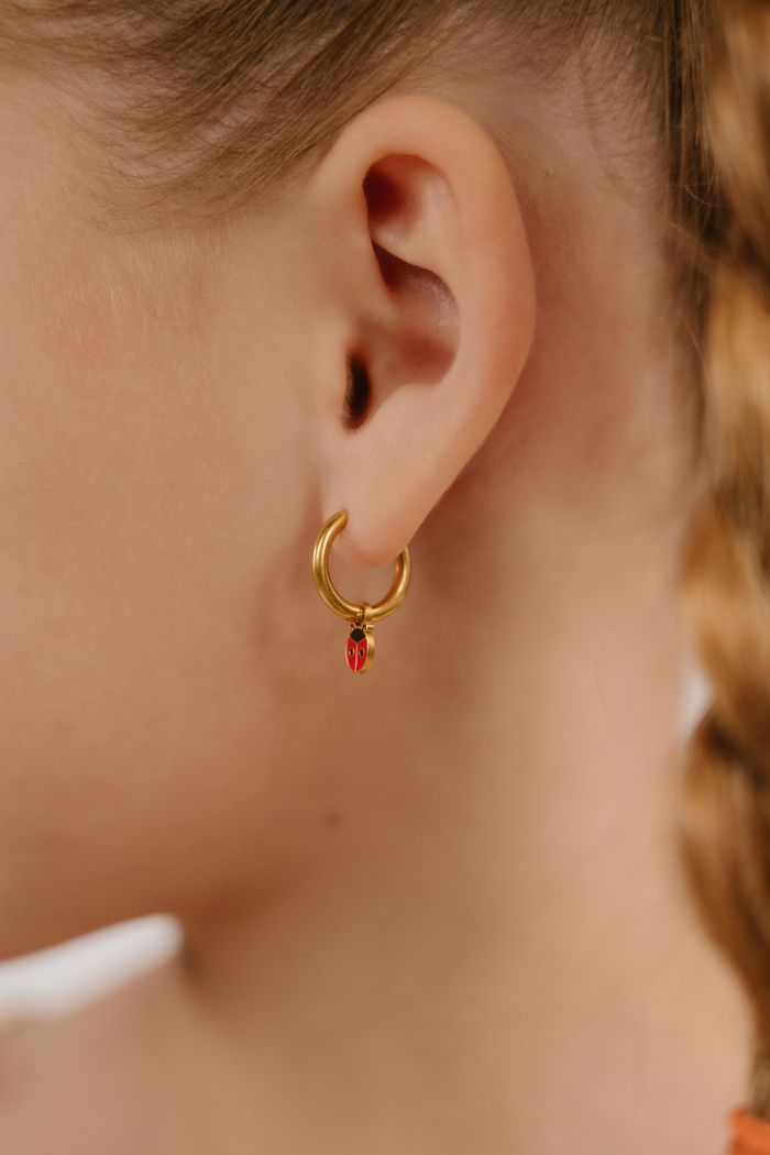 A close up of a ladybug shaped hoop charm with red and black enamel and gold-plated titanium metal hanging from a hoop earring.