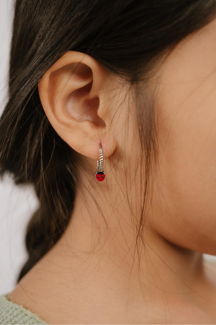 A close up of a ladybug shaped hoop charm with red and black enamel and  titanium metal hanging from a hoop earring.