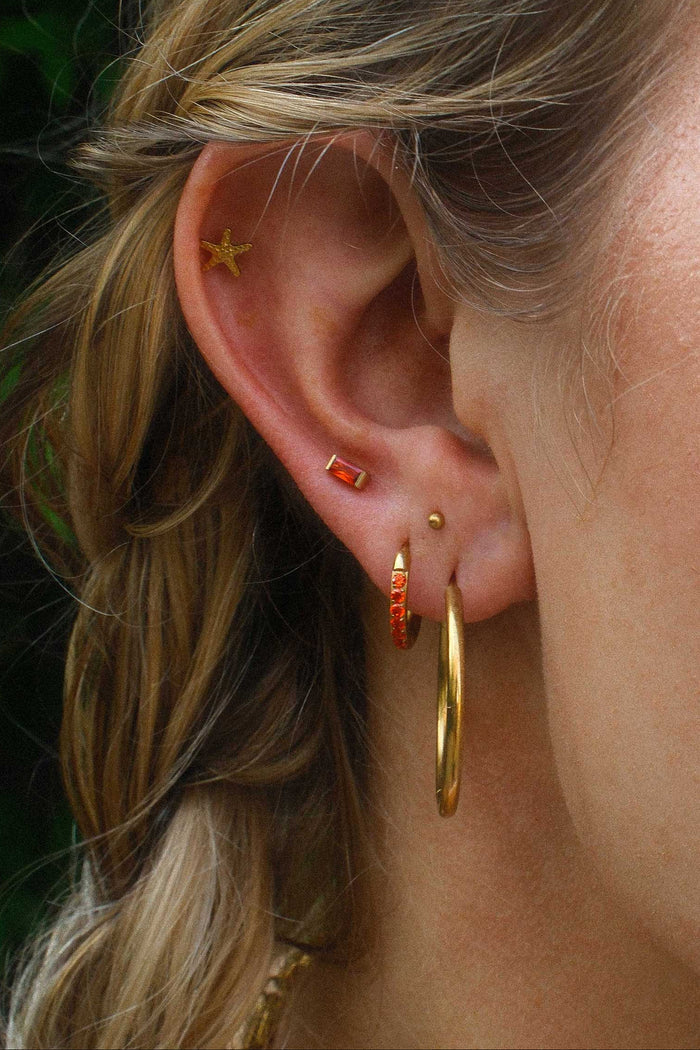 Close-up of an ear with gold hoop earrings and orange studs against a blurred background.