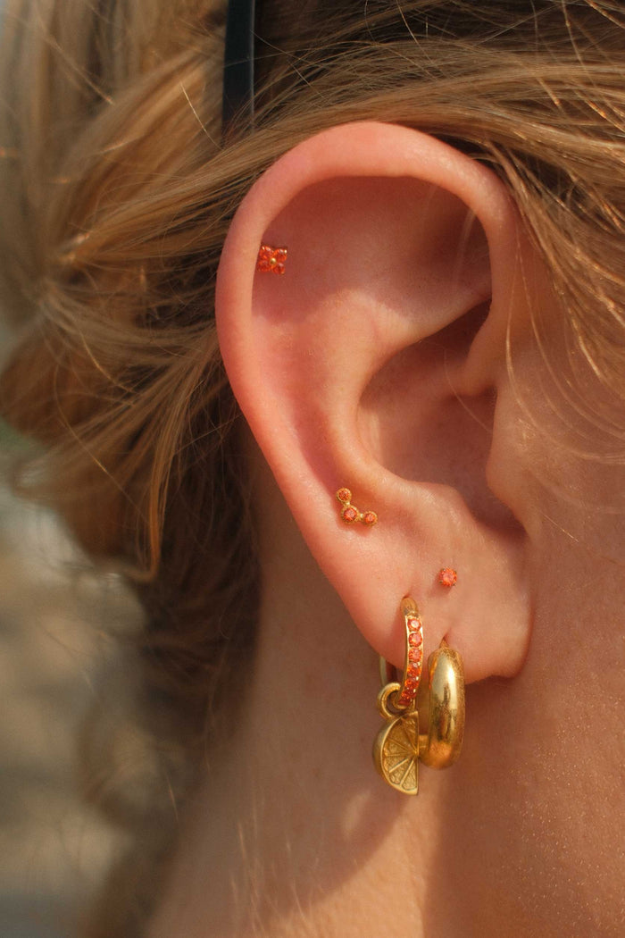 Close-up of an ear with gold earrings and small orange studs against a blurred natural background