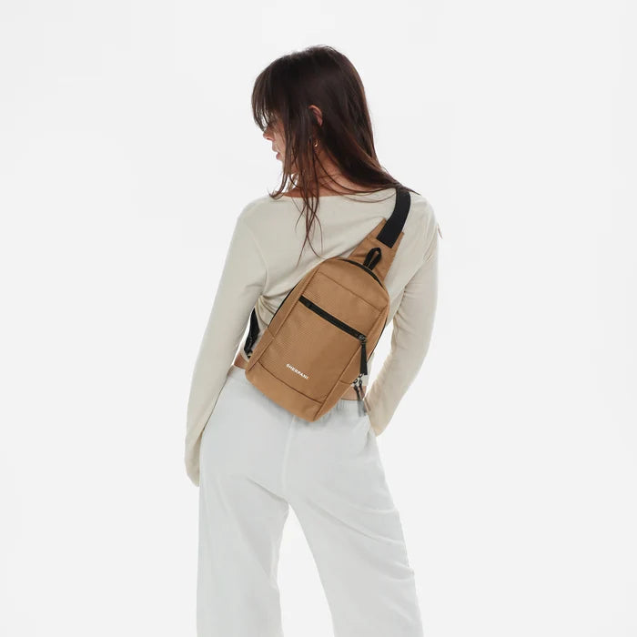 Woman wearing a Sherpani Metro Sand Anti-theft Reversible Sling on her back against white backdrop. #color_sand-tan