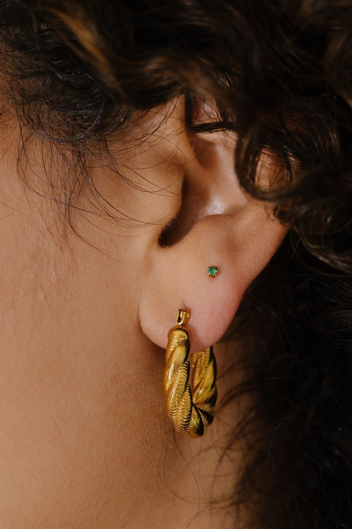 Close-up of an ear wearing a gold hoop earring and a tiny green CZ gemstone in the second lobe piercing.