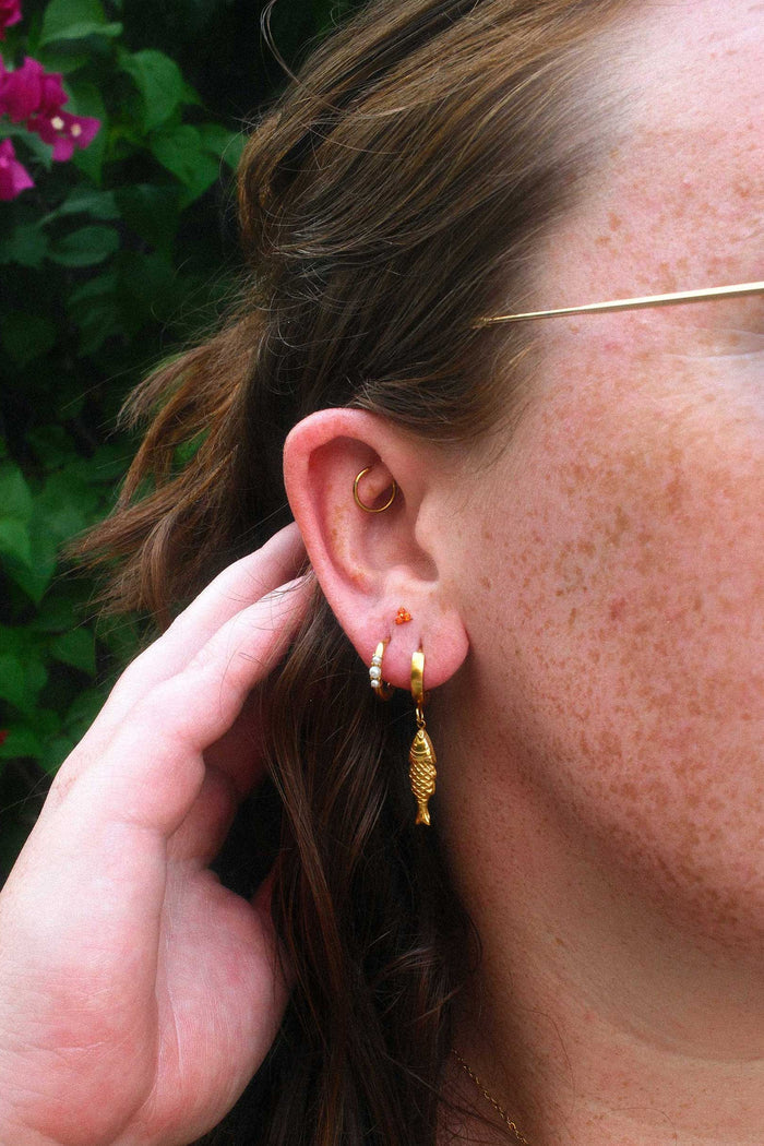 Close-up of a person wearing gold hoop earrings and an orange flat back stud with a blurred background
