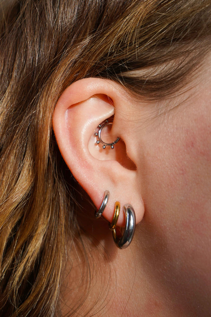 A close up of a model's ear wearing multiple earrings, including a hoop earring that features a split design where one half of the hoop is silver in color and the other is gold. The hoop is made from ASTM F136 Implant Grade Titanium and the gold half is finished with 18k Gold PVD plating.