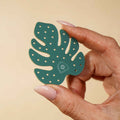 Hand holding a green leaf-shaped acrylic piece with little circle cutouts on a beige background