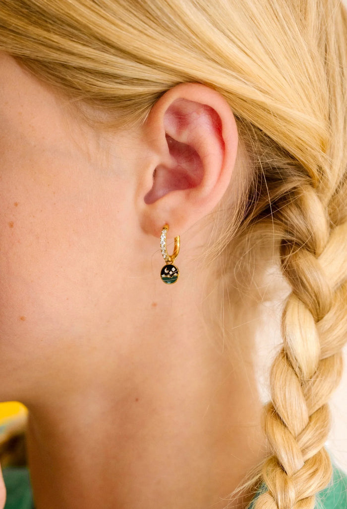 Close-up of an ear wearing a gold earring with a night sky scene made from enamel and clear gemstones, blonde hair in a braid.