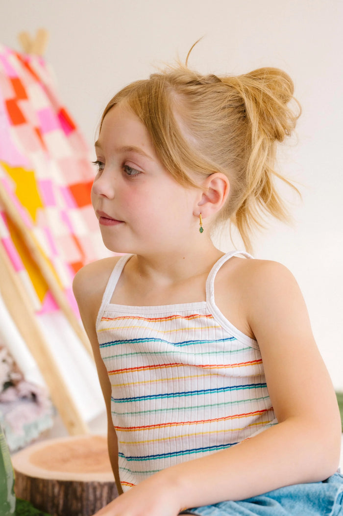 Young child sitting indoors wearing a colorful striped tank top and denim skirt.