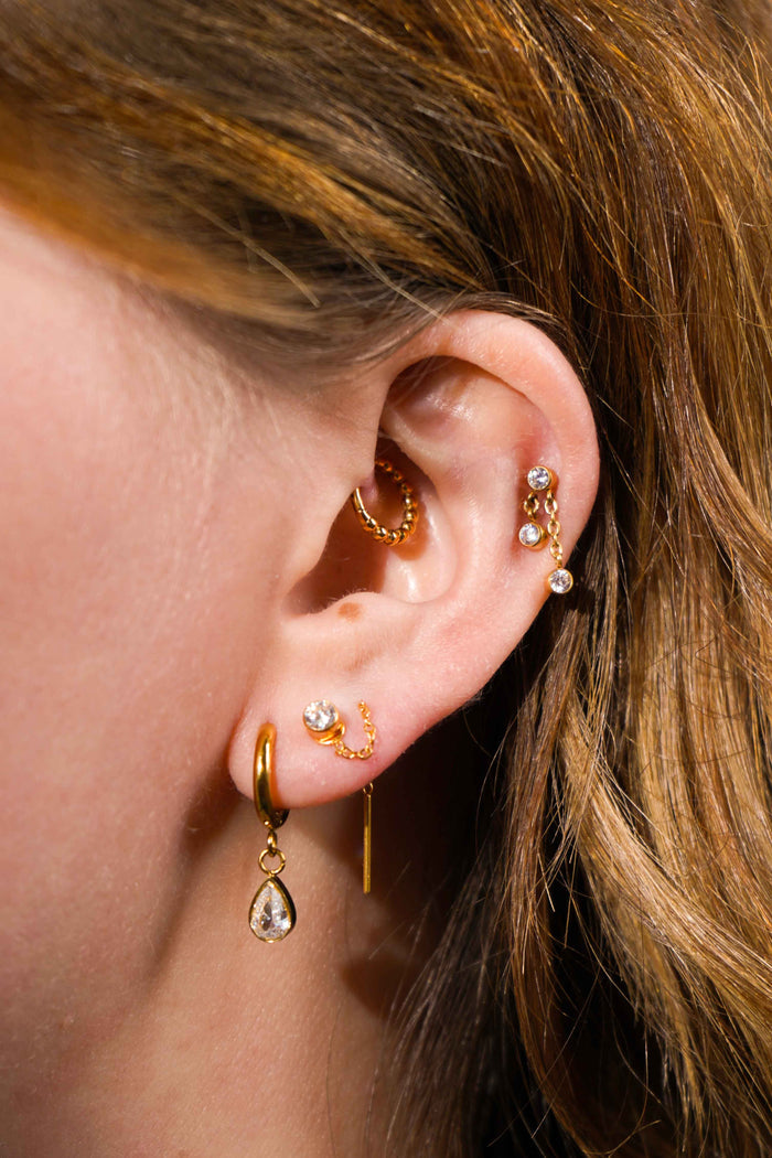 Close up of a model's ear, with multiple earrings including the featured product, a push-pin style flat back stud featuring a bezel-set round zirconia stone, clear in color, with 2 additional stones of the same style and color that hang from chains attached to the main stone. The flat back is made from ASTM F136 Implant Grade Titatnium, and is finished with 18k Gold PVD plating.