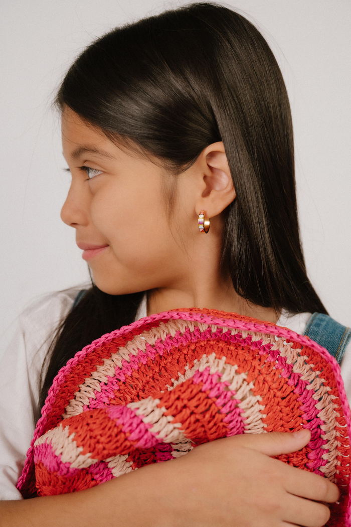 A  model wearing a hoop earring made from 18k gold-plated titanium metal, with geometric colored patterns that are reminiscent of quilt blocks in pinks, purple, blue, and green on a white background.