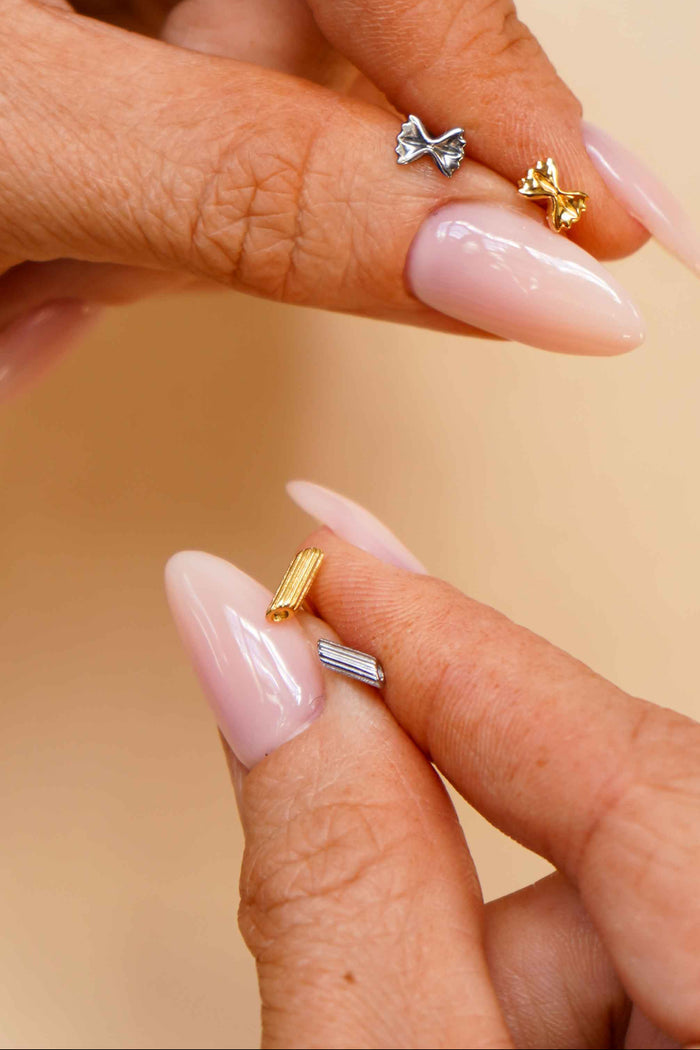 Close-up of a hand holding multiple gold and silver flat back stud earrings that are shaped like different pasta noodles.