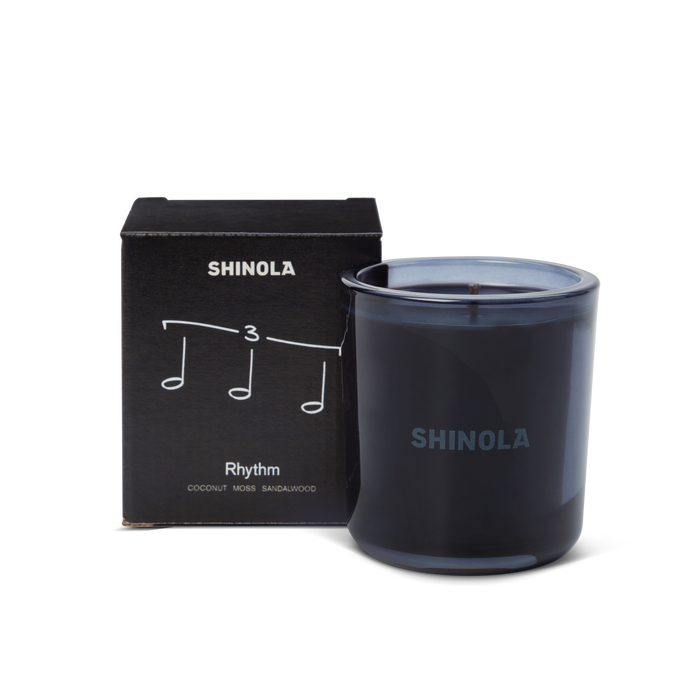 The Shinola Rhythm 8 oz Scented Candle sits next to its box decorated with musical notes and "Rhythm," inspired by Detroit nightlife. Scented with coconut, moss, and sandalwood, the candle comes in a recycled glass vessel.