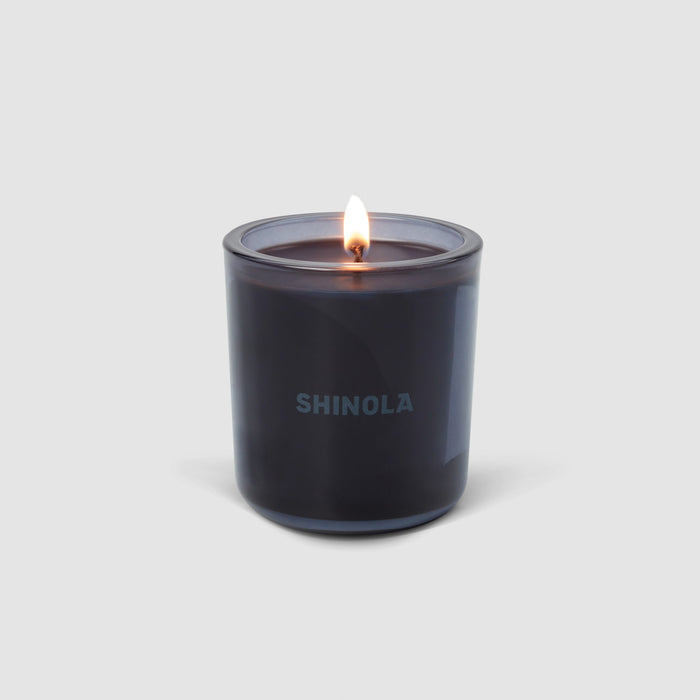 A Shinola Rhythm 8 oz Scented Candle glows warmly in a dark recycled glass vessel, “SHINOLA” printed on the front, set against a plain light gray background.