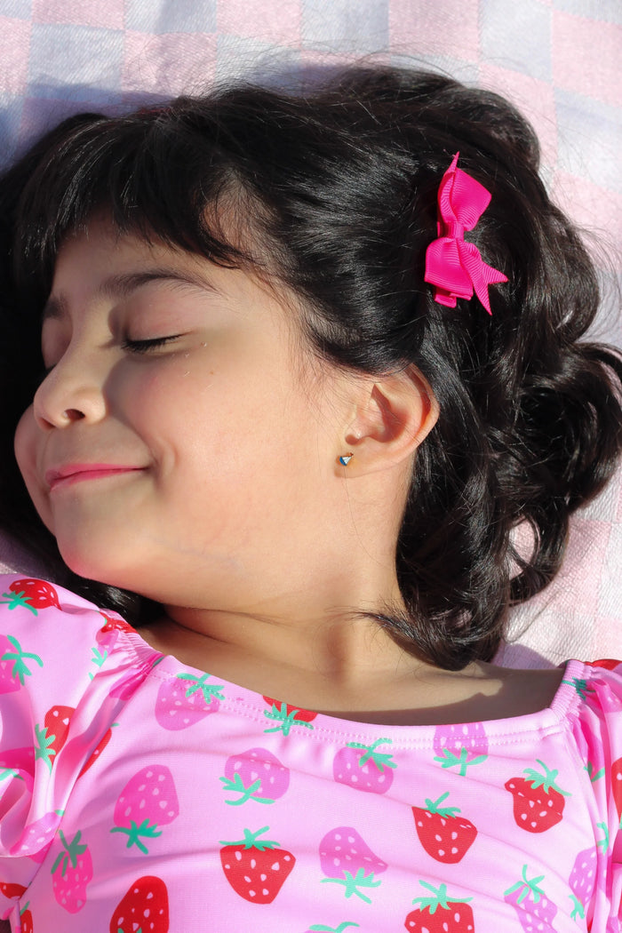 Young girl with a pink bow in her hair, wearing a pink shirt with strawberry pattern, lying on a checkered blanket.