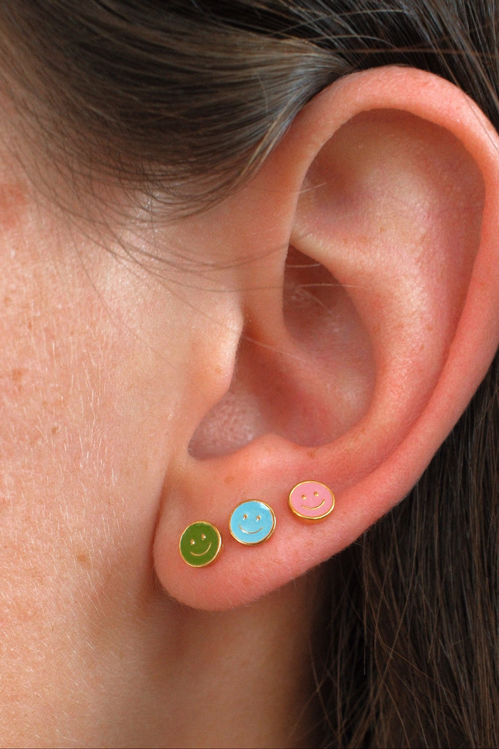 Close-up of a person's ear wearing three small smiley face stud earrings in green, light blue, and pink.