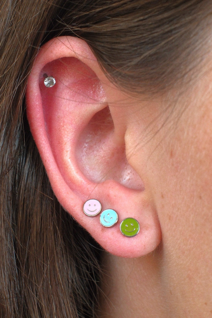 Close-up of a person's ear showcasing three colorful stud earrings: a pink smiley face stud, a light blue smiley face stud, and a lime green smiley face stud, with a small silver hoop earring in the upper ear cartilage.