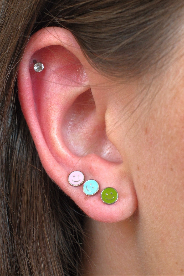 Close-up of a person's ear showcasing three colorful stud earrings: a pink smiley face stud, a light blue smiley face stud, and a lime green smiley face stud, with a small clear cz stud earring in the upper ear cartilage.