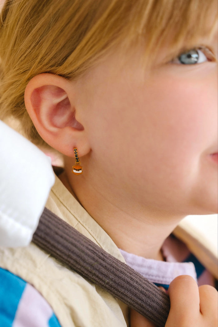 Close-up of a person wearing gold hoop earrings with enamel campfire charm, a red bandana, and a white shirt.