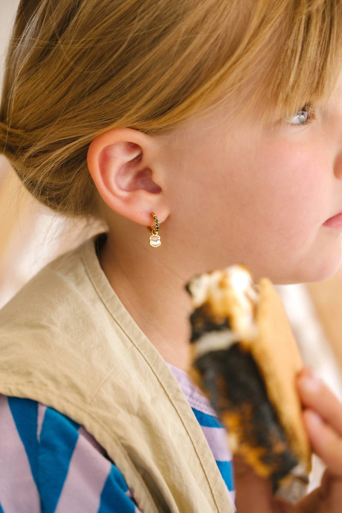 Close-up of a person wearing gold hoop earrings with enamel campfire charm, a red bandana, and a white shirt.