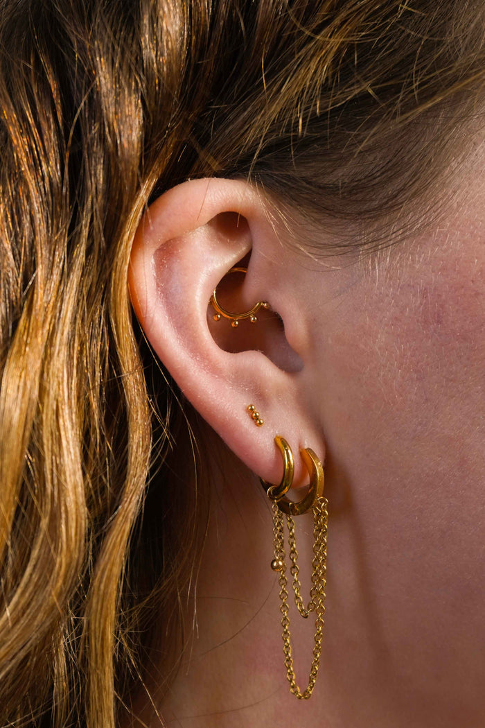 Close up of a model wearing multiple earrings, including clicker-style hoop featuring a series of round metal details that radiate from the main hoop in the Daith piercing. Made from ASTM F136 Implant-Grade Titanium, finished with 18K Gold PVD plating
