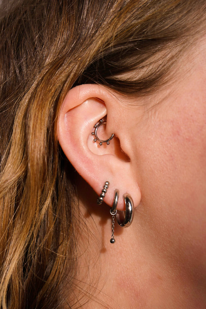 Close up of a model wearing multiple earrings, including clicker-style hoop featuring a series of round metal details that radiate from the main hoop in the Daith piercing. Made from ASTM F136 Implant-Grade Titanium