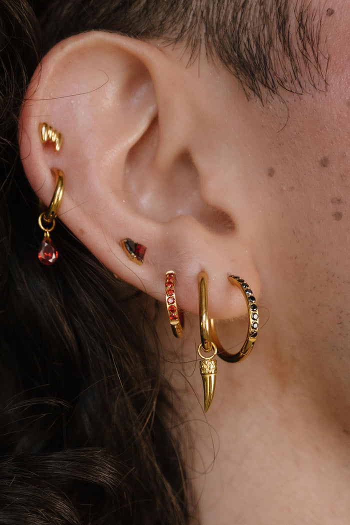 Close-up of a person's ear wearing multiple gold hoop and cartilage earrings, with small red gemstone accents and a gold leaf-shaped earring, against dark curly hair and a gold chain necklace.