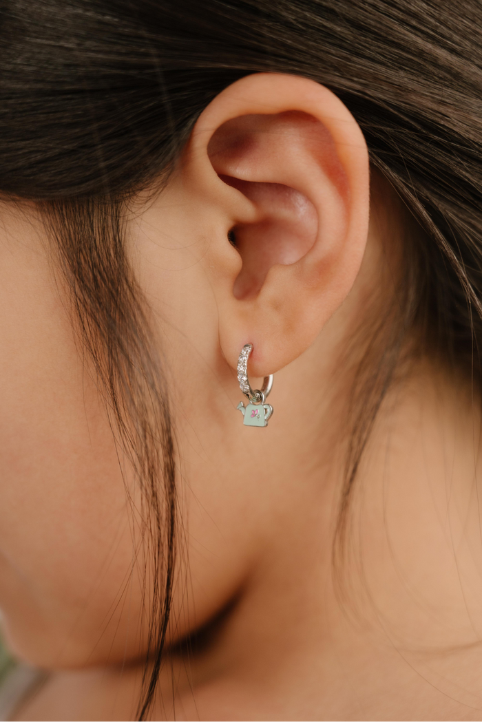 A close up of a right-facing watering can shaped charm with mint green enamel and titanium metal hanging from a hoop earring.